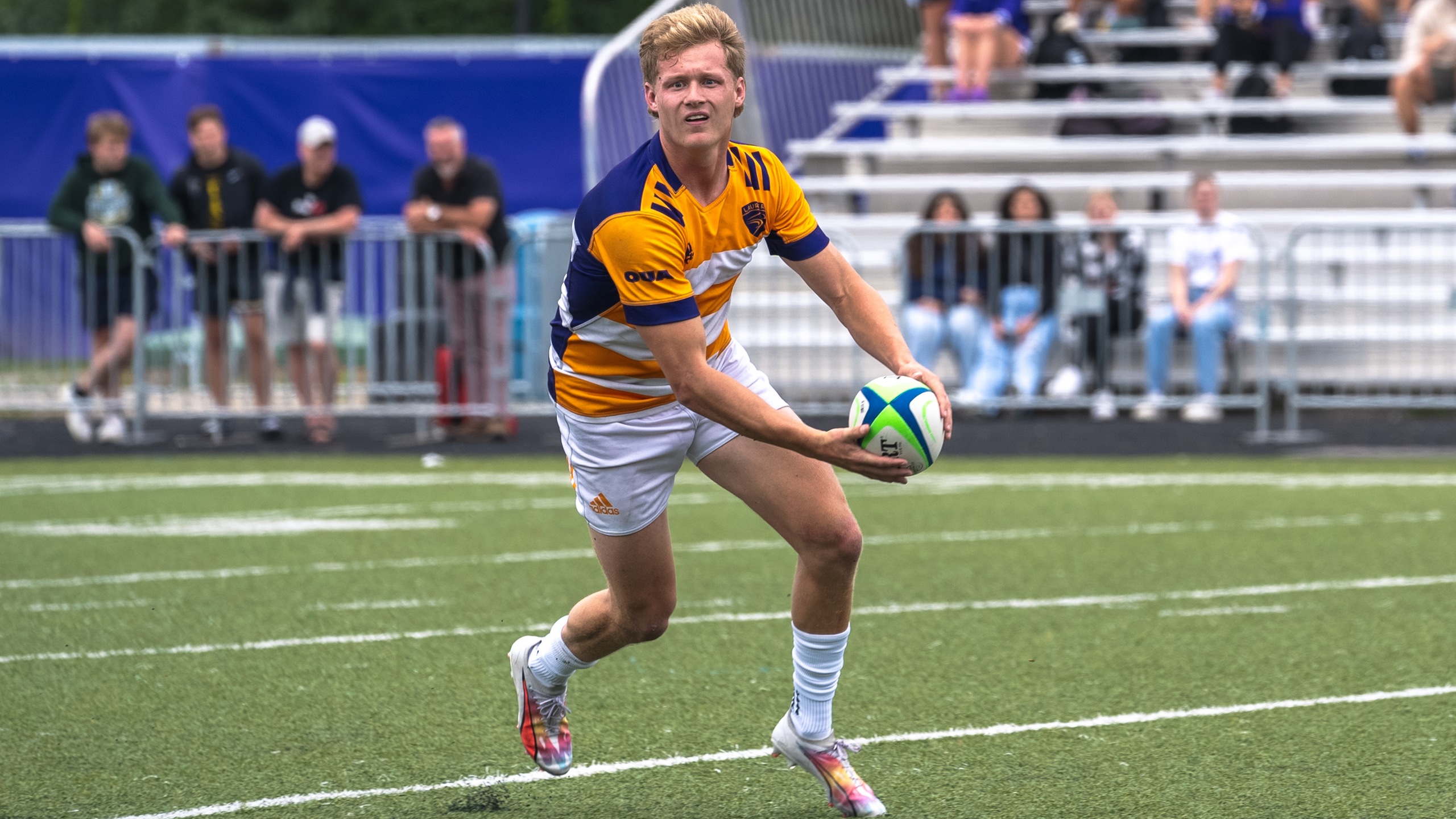 young man in golden yellow and navy blue soccer jersey and white shorts on a rugby pitch with a rugby ball in his hands getting ready to thrown.