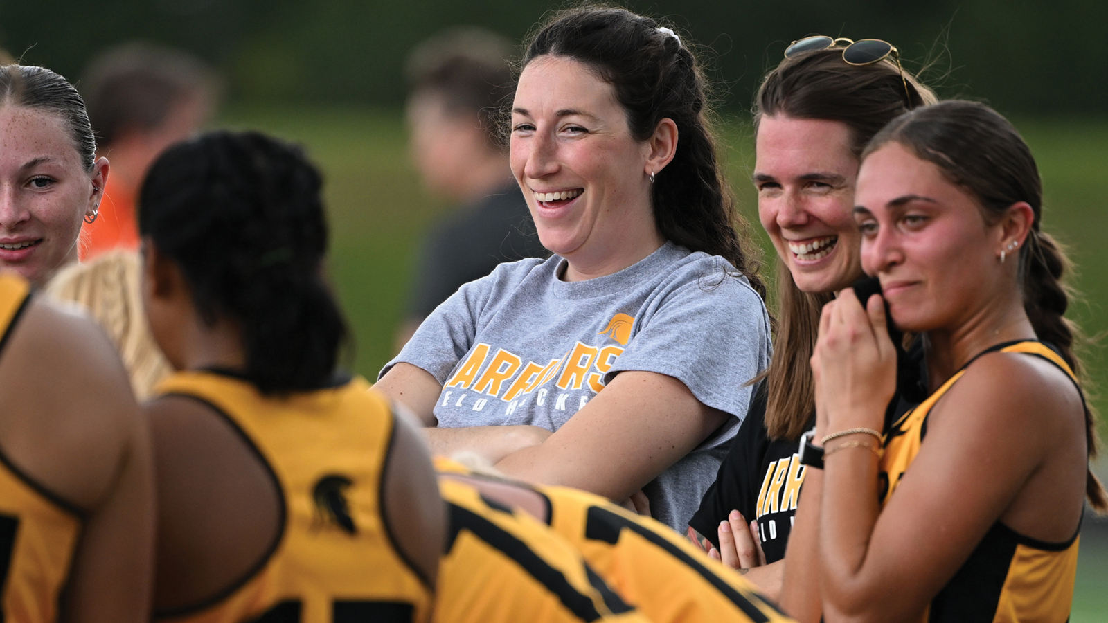 Action photo of Waterloo field hockey coach Maria Leahy laughing while part of a huddle with the Warriors team