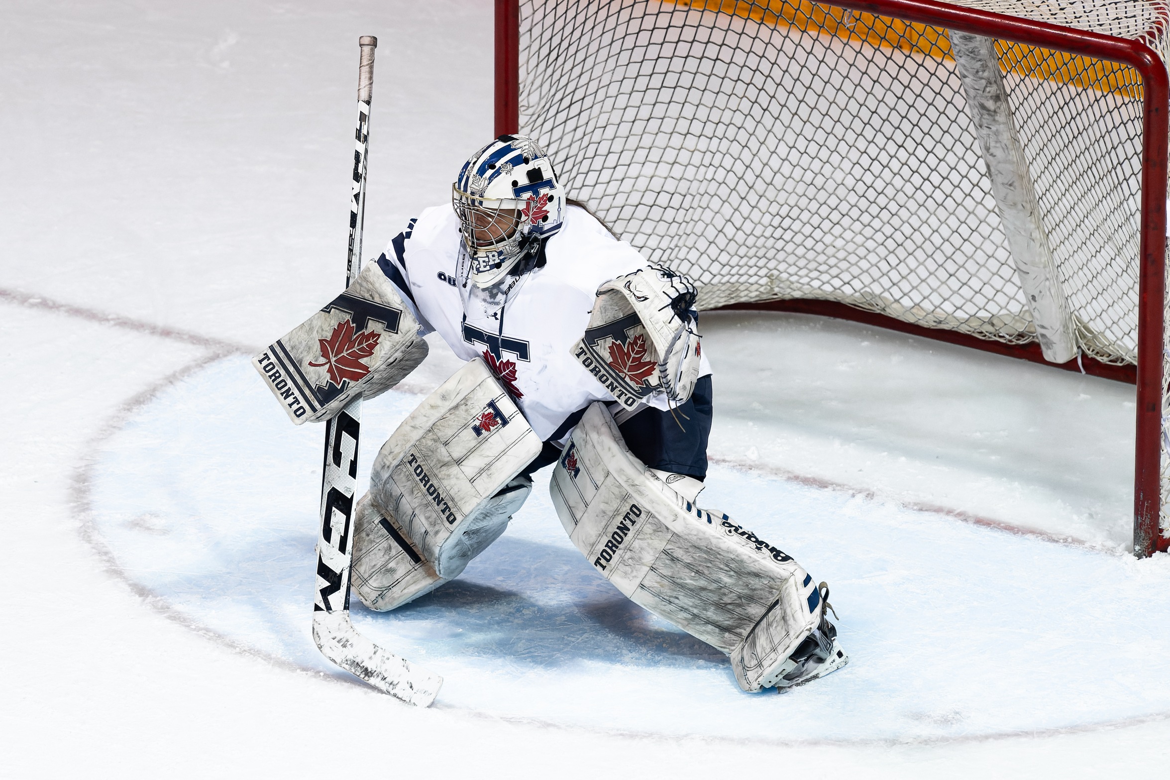 Image of women's hockey athlete in white gear and padding representing the Toronto Varsity Blues. Goalie is standing in front of net.