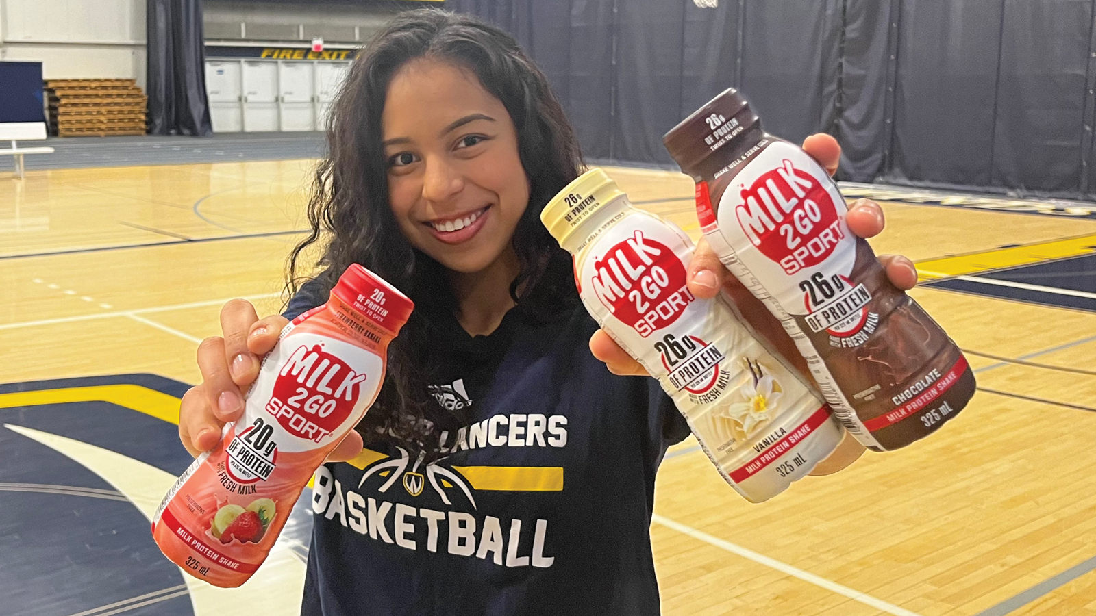 Windsor women's basketball player and 2023 Milk2Go Sport brand ambassador Isabella Anes standing in the middle of the school's basketball court, wearing a Windsor Lancers t-shirt and holding up three bottles of Milk2Go Sport in various flavours