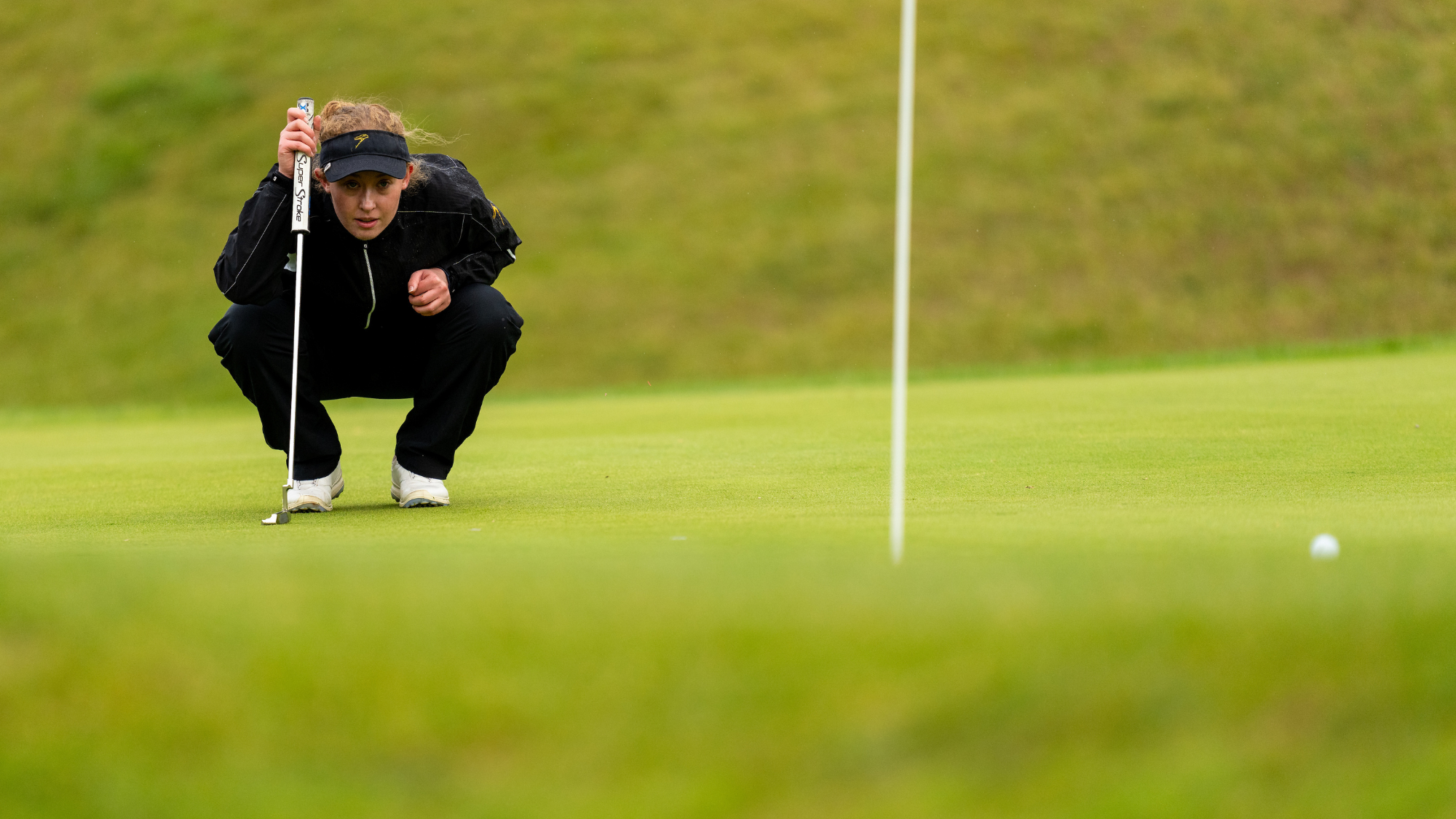 Woman in navy blue crouching on the golf course holding a golf club looking down and forward
