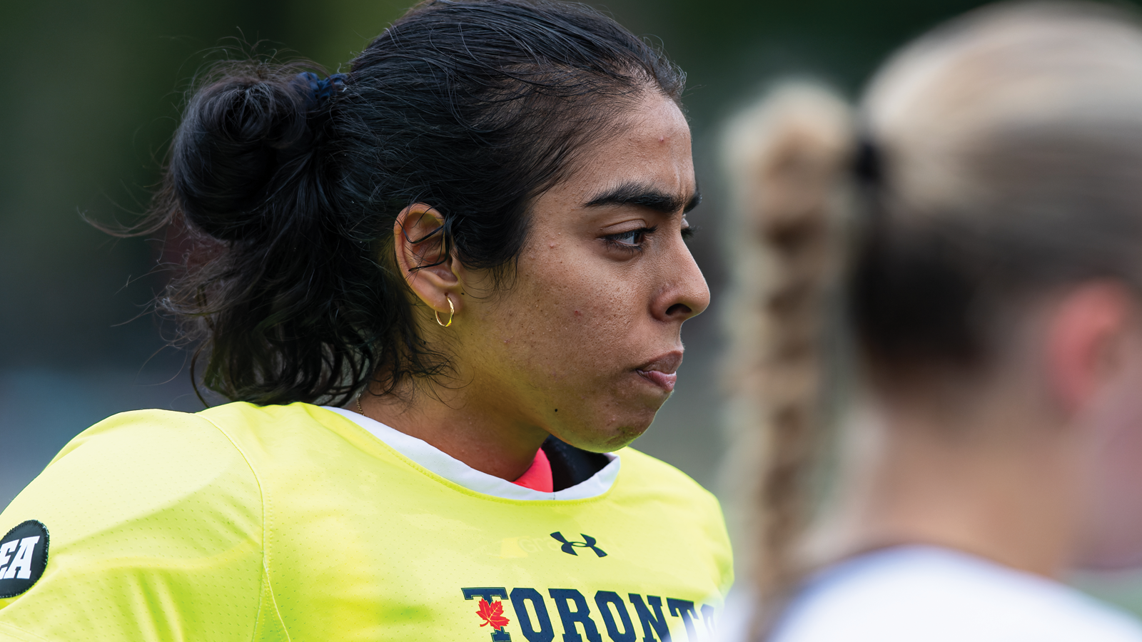 Close up of Toronto field hockey goalie Deepi Gill in in a bright yellow jersey staring in towards a team huddle