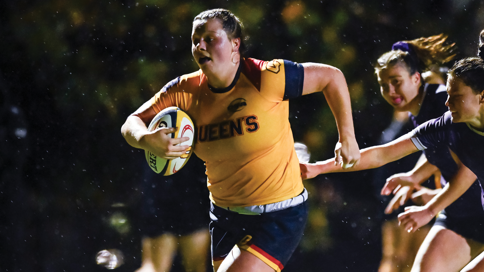 Queen's women's rugby player Maggie Banks running with the ball tucked in to her right arm as opposing players try to tackle her from behind