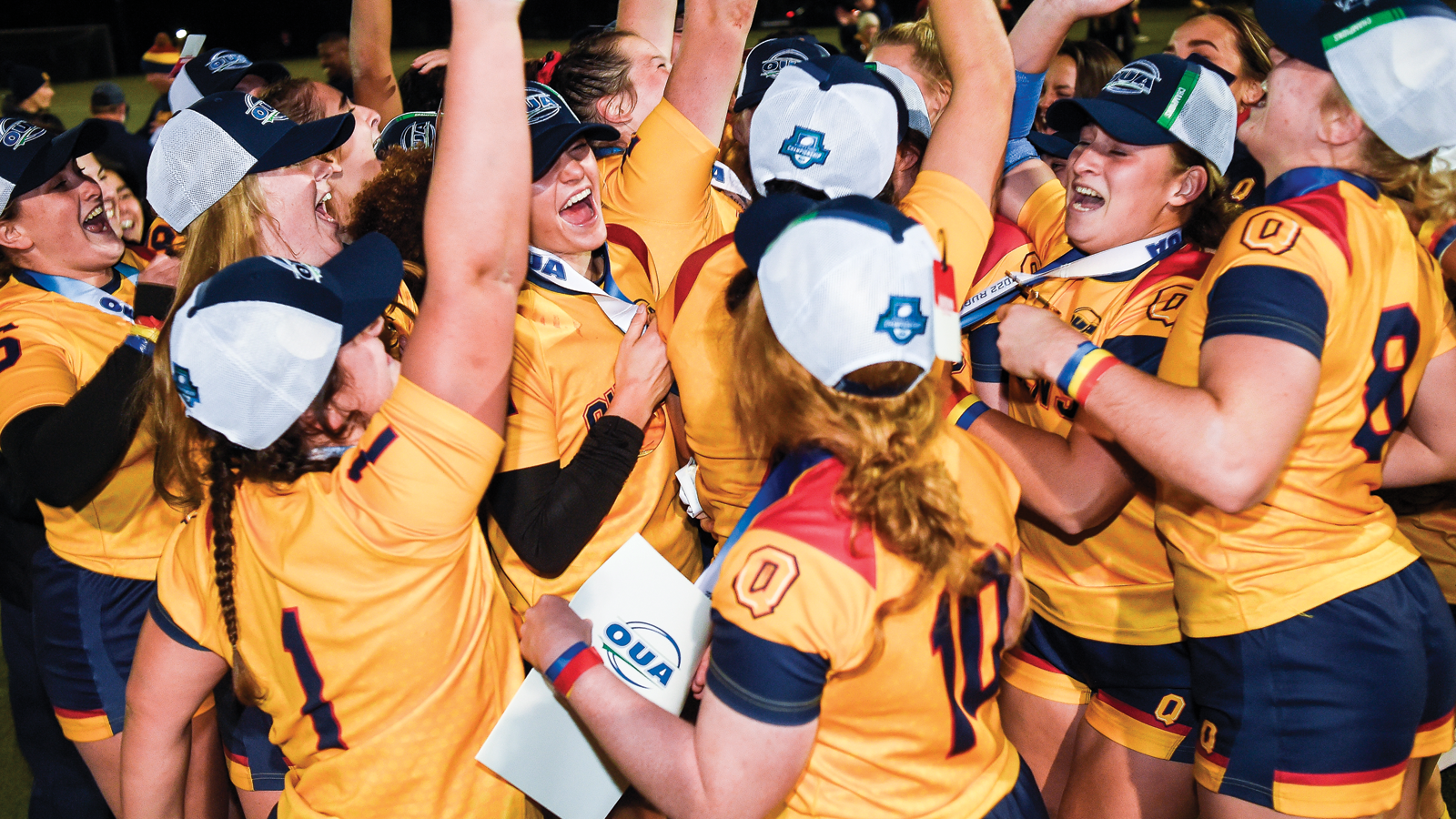 Queen's women's rugby team celebrating in a huddle post-game, wearing OUA champion hats and wearing medals around their neck