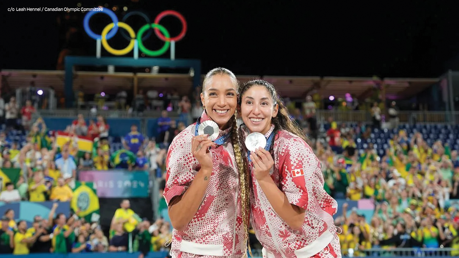 Beach volleyball players Melissa Humana-Paredes and Brandie Wilkerson posing with the 2024 Olympics silver medals around their neck, with the crowd and Olympic rings in the background