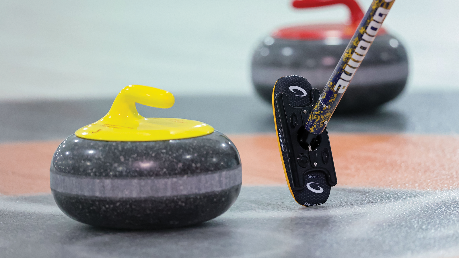 Two curling rocks on the ice with a broom angled beside the yellow stone in the foreground