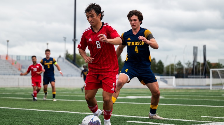 Action photo of York men's soccer player with the ball at his feet as a Windsor player approaches to defend