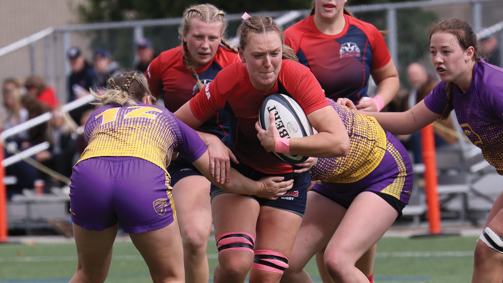 Action photo of Brock women's rugby player Lily Freiburger running with the ball during a game while being tackled by two opposing Laurier players