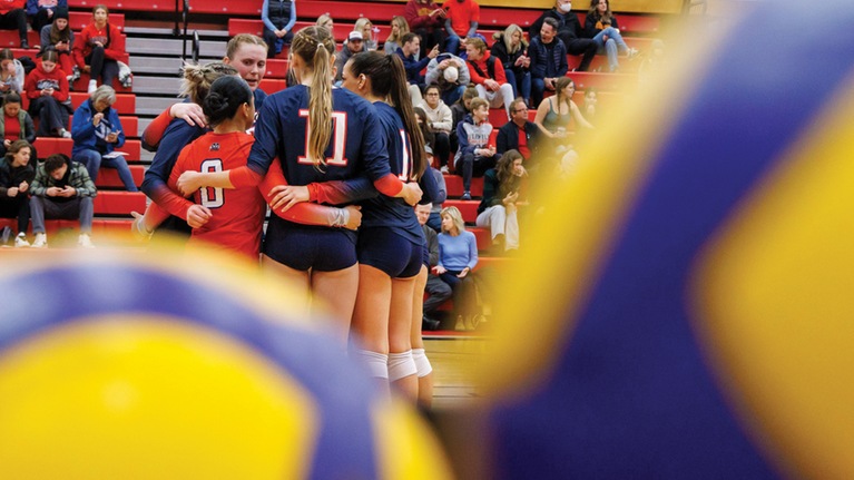 Brock women's volleyball team huddled together on the court in the background, with two blurred volleyballs showcased in the foreground