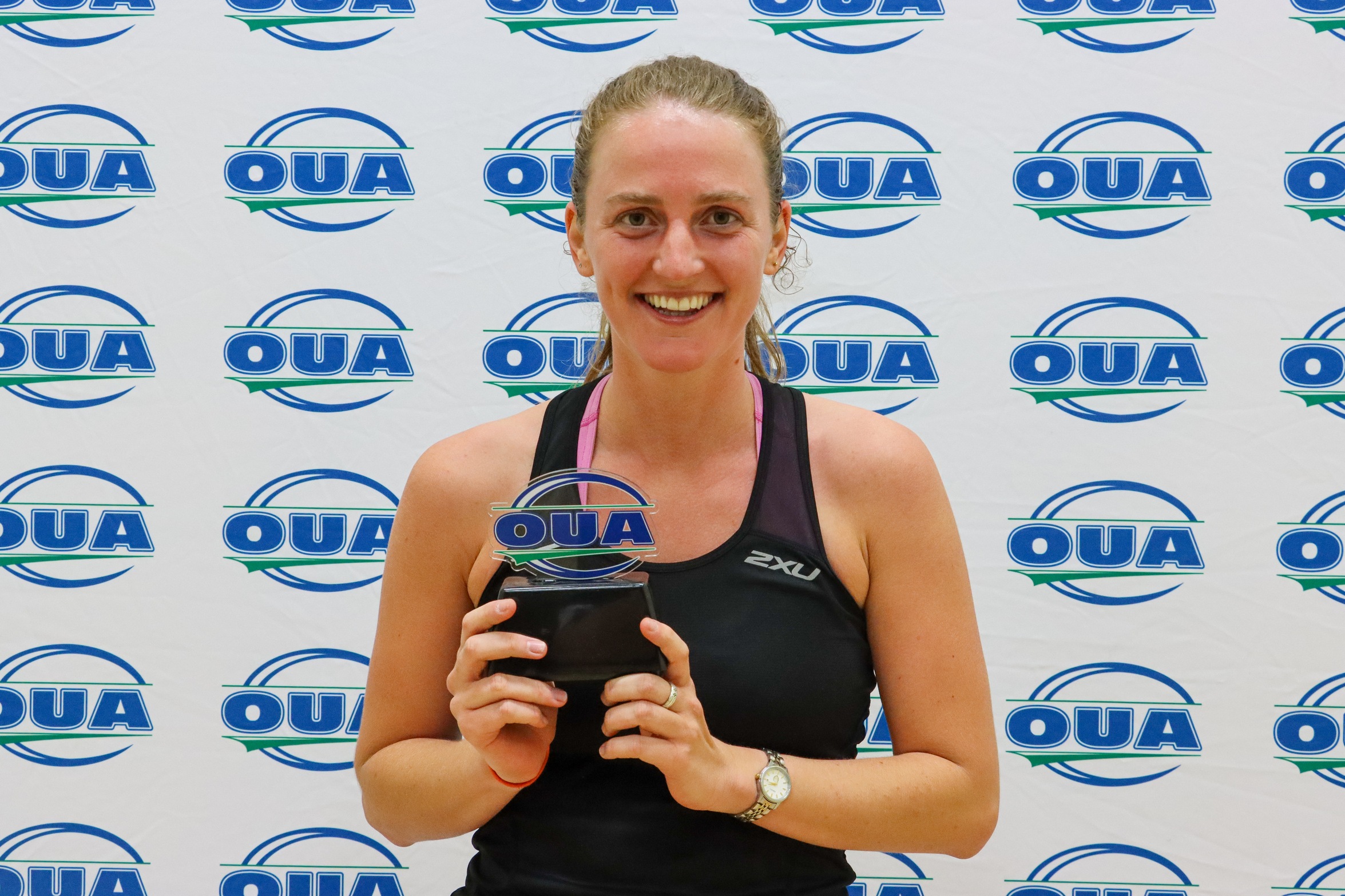Queen's women's squash player Maddie O'Connor posing in front of an OUA step-and-repeat backdrop holding an OUA trophy